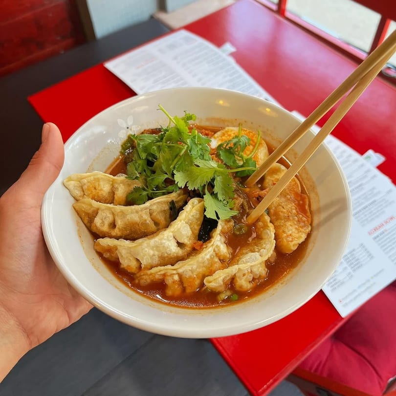 A hand holding a bowl of Kameya Ramen & Sushi's gyoza in broth with cilantro and chopsticks.